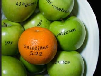Green apples in a bowl with one orange labeled 'Galatians 5:22', symbolizing the fruits of the Spirit.